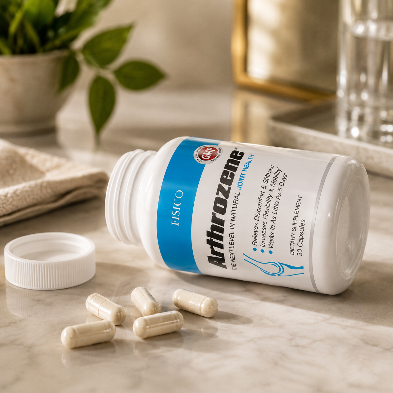 A white bottle of FSISCO ArthroZenie joint health capsules lies on its side on a light marble surface, with several capsules scattered in front and the white cap placed beside it. The background is softly blurred with a plant and glassware.