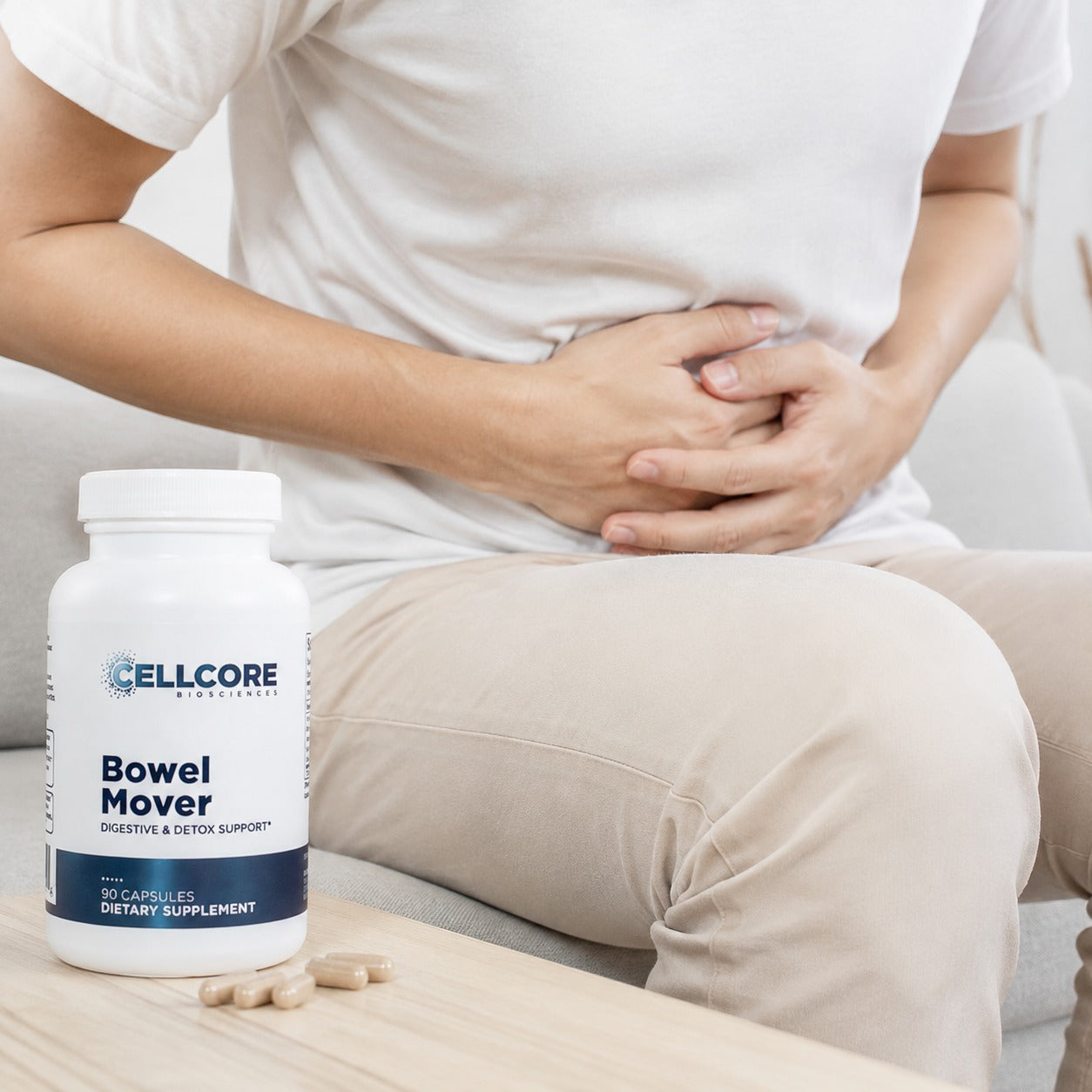 A person sitting on a couch holding their stomach next to a white bottle of  Bowel Mover dietary supplement with a few capsules on a wooden table.